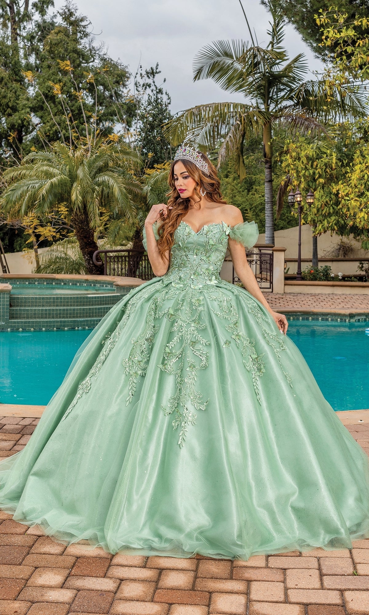 A woman wearing the Quinceanera Dress 1834 By Dancing Queen, featuring off-the-shoulder floral embroidery and a sweetheart neckline, stands by a pool among palm trees, adorned with a tiara.